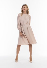 Rochie cu mânecă lungă, de culoare roz pal, cu decolteu rotund, talie strânsă și fustă fluidă. Prezintă manșete delicate cu plisaje și o curea legată.