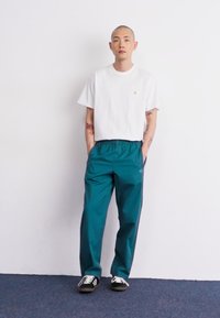 T-shirt blanc à manches courtes avec un col rond, associé à un pantalon élastique taille haute teal. Des baskets noir et blanc complètent la tenue.