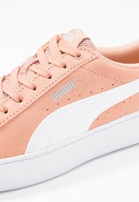Jasnołososiowe zamszowe sneakersy z białymi akcentami, teksturowanym siateczkowym językiem, płaskim zapięciem na sznurowadła oraz logo Puma w metalicznie srebrnym kolorze po boku.