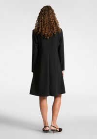 Schwarzes A-Linien-Kleid mit langen Ärmeln, das ein schlichtes Design, eine glatte Textur und einen knielangen Saum aufweist. Dazu werden braune Plateausandalen getragen.