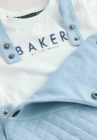 Bela majica s plavim logotipom "BAKER" in svetlo modri podloženi kombinezon z kovinskimi pritiskači, ki ima ravne naramnice in teksturirano tkanino.