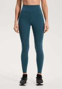 Magas derekú, türkizkék leggings elasztikus anyagból, sima textúrával és díszítő varrásokkal, fekete sportcipővel párosítva.