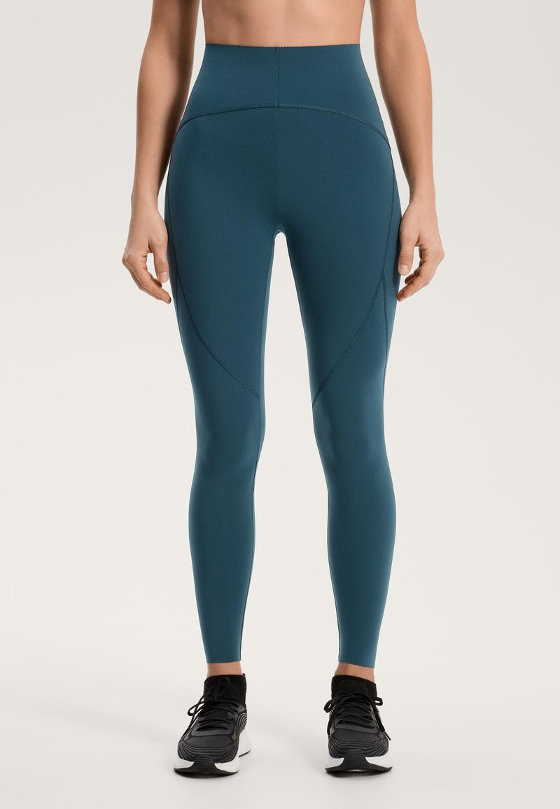 Magas derekú, türkizkék leggings elasztikus anyagból, sima textúrával és díszítő varrásokkal, fekete sportcipővel párosítva.