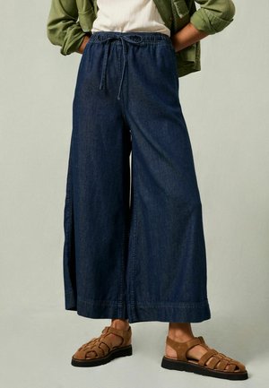 Denim wide-leg pants in dark blue with an elastic waistband and drawstring. Featured with side slits for added movement. Paired with brown sandals.