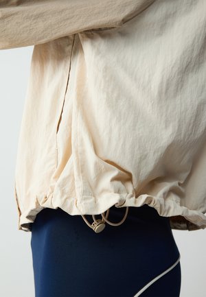 Lichtbeige jas met een gekreukeld textuur, verstelbare trekkoord onderrand, gecombineerd met aansluitende donkerblauwe broek met een contrasterende witte bies.