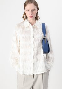 Blusa bianca trasparente con texture a zigzag, dotata di colletto e dettagli sfilacciati. Abbinata a una borsa a tracolla blu.