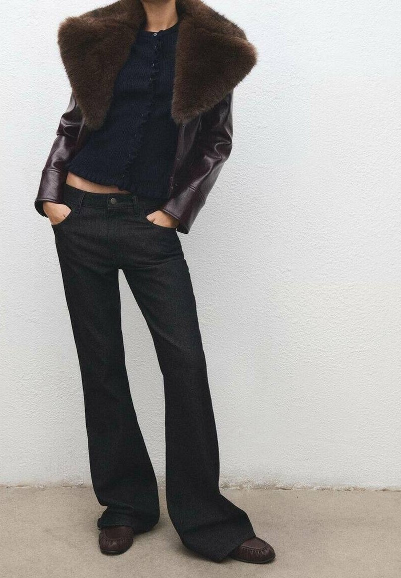 Personne portant une veste en cuir marron foncé avec un grand col en fourrure, un haut noir boutonné, un pantalon large noir et des chaussures marron foncé, les mains dans les poches.