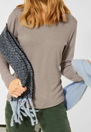 Persoon die een beige overhemd met lange mouwen en een groene broek draagt, en een blauw-zwart geruit sjaal met franjes vasthoudt.