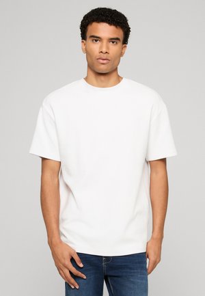 Ung mand med kort krøllet hår iført en ensfarvet hvid tekstureret kortærmet skjorte og blå jeans, stående mod en grå baggrund.