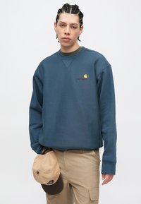 Modrá mikina s kulatým výstřihem vyrobená z měkké látky, ozdobená malým žlutým a červeným logem. Doplňuje ji béžová baseballová čepice držená v ruce.