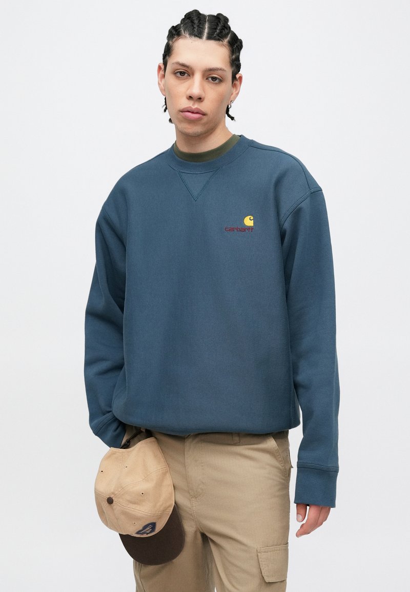 Blå crewneck-sweatshirt lavet af blødt stof, med en lille gul og rød logo. Kompletteret med en beige baseballkasket i hånden.