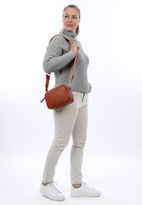 Vrouw die een grijze coltrui, lichte broek en witte sneakers draagt, met een bruine leren schoudertas over haar schouder, kijkt zijwaarts.