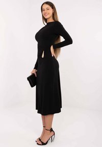 Czarna sukienka z długim rękawem, dopasowanym gorsetem i rozszerzaną spódnicą. Posiada marszczenie z boku z złotym akcentem. Modelka trzyma czarną kopertówkę.