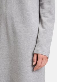 Abito grigio a maglia con maniche lunghe, caratterizzato da una texture morbida e un orlo con polsini. Una mano poggia sul tessuto, mostrando la vestibilità rilassata.