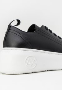 Gros plan sur une basket noire avec une semelle blanche épaisse et un logo circulaire en relief sur le côté, avec une languette au talon et des lacets noirs.