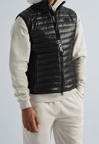 Gilet rembourré noir avec des coutures verticales et une capuche. Il dispose de deux poches latérales zippées et d'un zip avec logo. Porté par-dessus un sweat-shirt gris clair.