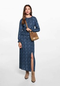 Temno modra obleka z dolgimi rokavi in belim geometrijskim vzorcem, z razporkom na strani; v kombinaciji z rjavo torbico in bež gležnjarji.