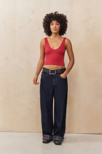 Top corto rosso a coste, jeans a gamba larga blu scuro con risvolti a quadri, cintura nera con una grande fibbia, indossato con scarpe nere, su uno sfondo in legno chiaro.