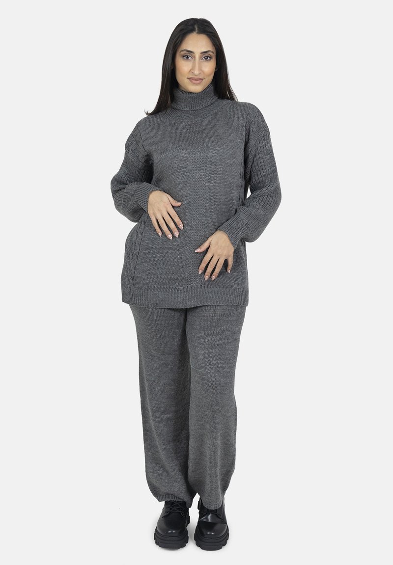 Grå strikket sweater med høj krave og teksturerede ærmer, parret med løstsiddende grå bukser og sorte kraftige sko.