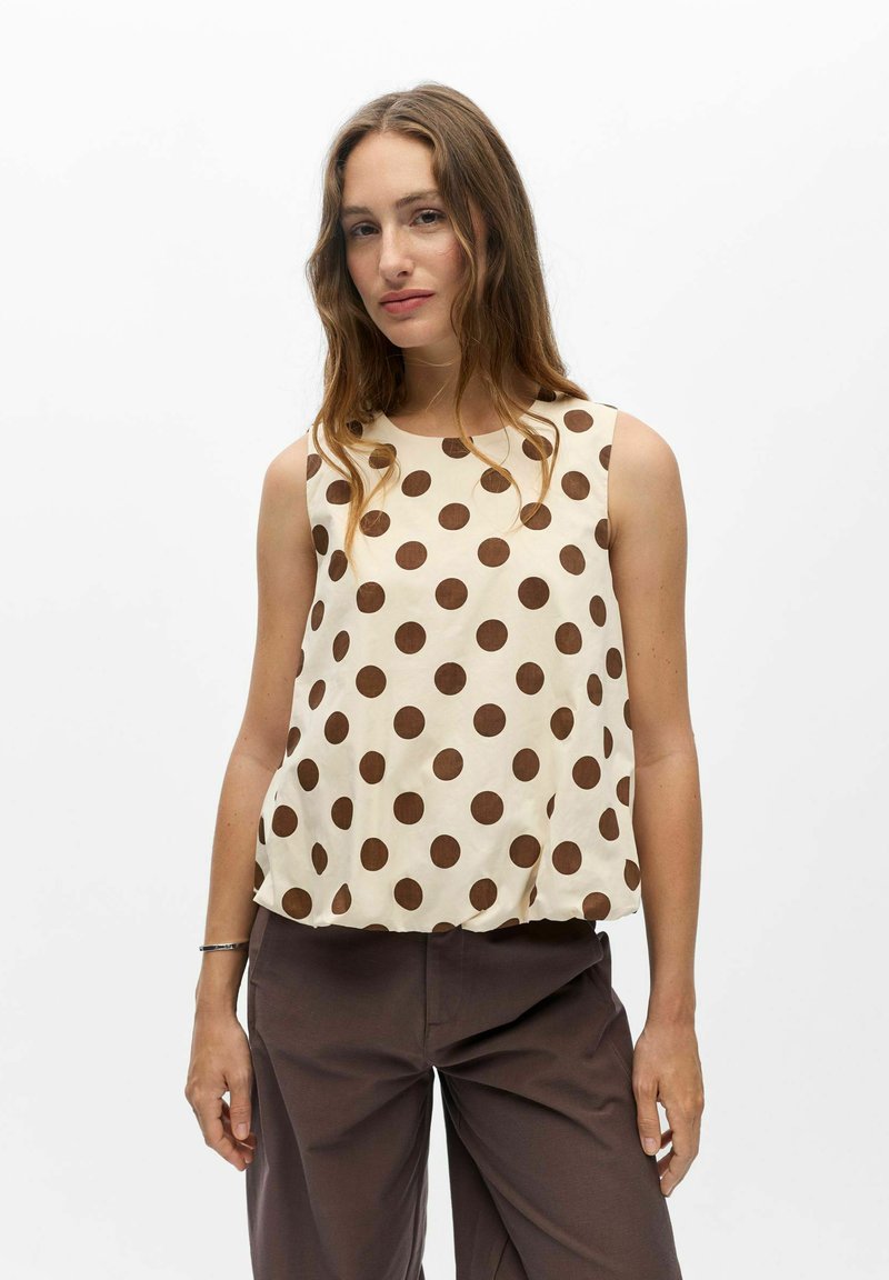 Femme aux longs cheveux bruns portant un haut beige sans manches à gros pois marron et un pantalon marron foncé sur fond blanc.
