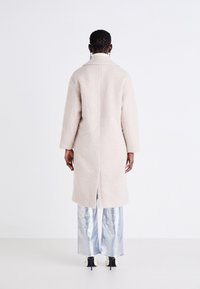 Cappotto beige strutturato con taglio dritto, colletto a scialle e spacchetto centrale sul retro, abbinato a pantaloni ampi argentati lucidi e tacchi.
