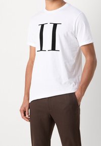 Camiseta blanca de algodón de manga corta, con un gran gráfico negro "II" en el centro. Combinada con pantalones marrón oscuro.