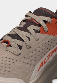 Κοντινή λήψη μπεζ και πορτοκαλί παπουτσιού για τρέξιμο με υφασμένα κορδόνια και διάτρητη πλευρική επιφάνεια με το λογότυπο "Altra" στην σόλα.