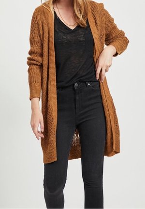 Femme portant un cardigan en tricot marron sur un haut noir semi-transparent à col en V et un jean noir taille haute, posant avec une main sur la hanche.