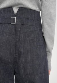 Pantalon taillé gris foncé en tissu texturé, doté d'une boucle arrière distinctive et de plusieurs poches, associé à un pull gris clair.
