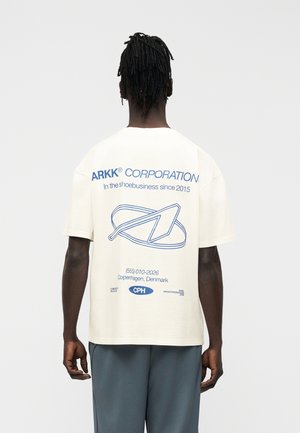 Άνδρας με σκούρο δέρμα και ρίζες στέκεται με την πλάτη γυρισμένη, φοράει λευκό T-shirt με μπλε κείμενο και γραφικό με βέλος και κύκλο.