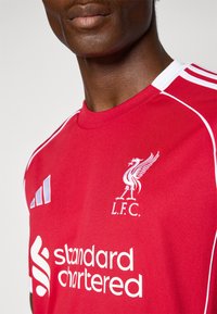 Tricou de fotbal roșu cu accente albe. Prezintă un logo brodat, „L.F.C.”, și textul „Standard Chartered”. Fabricat dintr-un material neted.