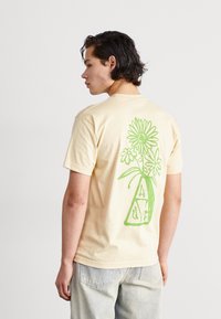 Camiseta de manga corta en color beige que presenta un gran diseño floral verde en la parte posterior, con un jarrón y letras estilizadas A, L, V, E.