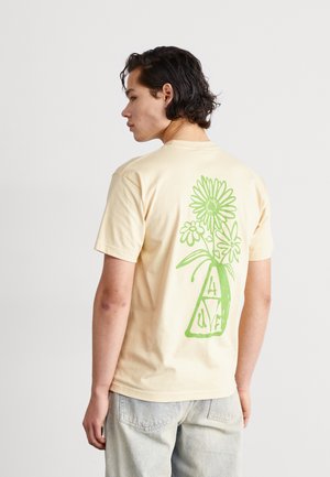 Camiseta de manga corta en color beige que presenta un gran diseño floral verde en la parte posterior, con un jarrón y letras estilizadas A, L, V, E.