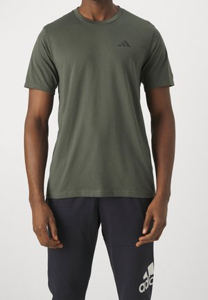 Mężczyzna ubrany w oliwkowo-zieloną koszulkę na krótki rękaw marki Adidas oraz czarne spodnie dresowe Adidas, stojący na jednolitym białym tle.