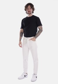 Pantaloni bianchi a vestibilità slim, abbinati a una t-shirt nera a maniche corte. Il modello indossa sneakers bianche con dettagli neri.