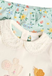 Weißes Baumwoll-Babytop mit gestickter Maus und Schmetterling, ausgestattet mit einem Spitzenkragen. Blaue Hosen mit floralem Muster, die dazu passen.