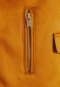 Tessuto scamosciato arancione con cerniera verticale, dotato di un tirante metallico e una tasca a pattina accanto alla cerniera. Texture liscia.