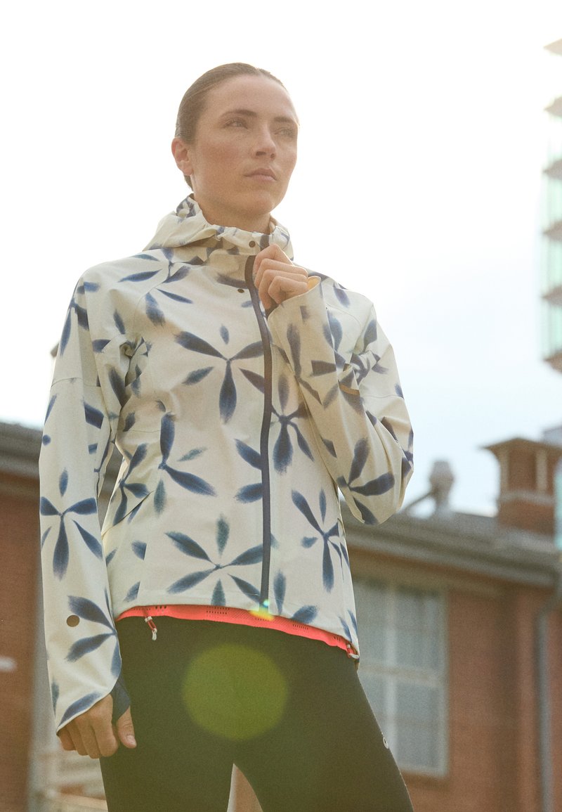 Leichtgewichtige Jacke mit Blumenmuster in Creme mit blauen Akzenten, mit Reißverschluss, Kapuze und belüftetem Saum, über schwarzen Leggings getragen.