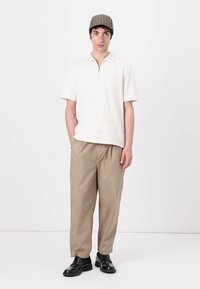 White short-sleeve polo with a zipper, beige relaxed-fit trousers, and black shoes. The model wears a plaid cap and stands against a plain background.