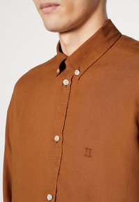 Camicia button-up marrone, con texture, dotata di colletto, bottoni bianchi e un dettaglio ricamato "II" sul lato sinistro del petto.
