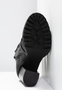 Botte en cuir noir levée contre un mur blanc, montrant un motif détaillé de la semelle et un talon bloc bas.