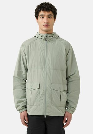 Joven de frente vistiendo una chaqueta con capucha color verde salvia claro, cremallera frontal y bolsillos con solapa, posando contra un fondo liso.