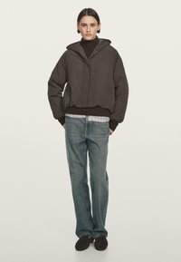 Persona che indossa un giubbotto imbottito con cappuccio scuro, un maglione a collo alto marrone, una camicia a quadri, jeans azzurri chiari e scarpe marroni scure, in piedi contro uno sfondo neutro.