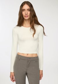 Crop top bianco a maniche lunghe realizzato in materiale morbido; presenta un collo rotondo e un design aderente. Abbinato a pantaloni attillati grigi.