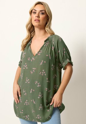 Mujer con cabello largo y rubio lleva una blusa verde con flores y mangas cortas, de pie frente a un fondo liso y claro.