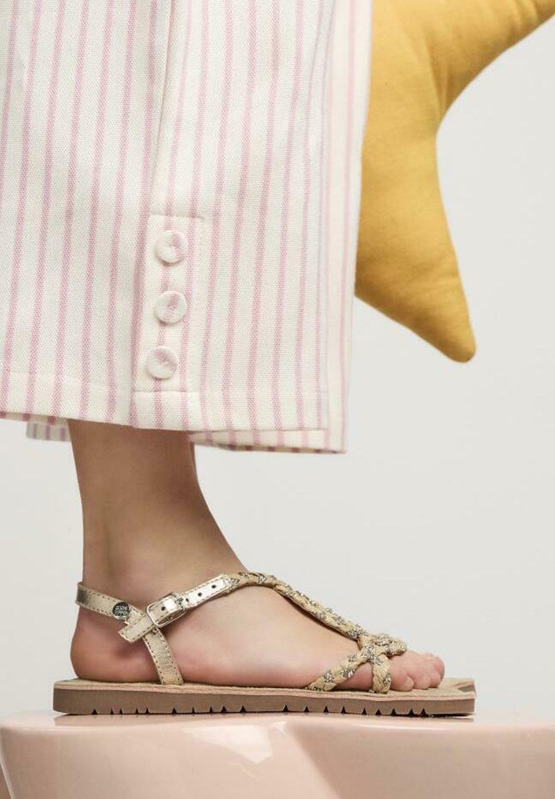 Sandalias doradas con tiras y detalles trenzados, con cierre de hebilla. El pie está posicionado sobre una plataforma rosa, con pantalones de rayas blancas y rosas visibles.