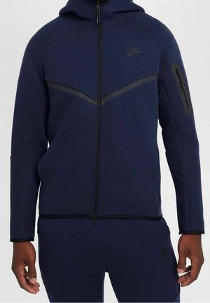Homme portant un sweat à capuche zippé Nike bleu marine foncé et un pantalon assorti avec une poche zippée sur la manche gauche, debout devant un fond uni.