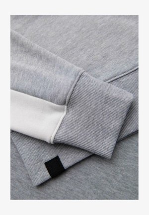 Hellgrauer Sweatshirt mit geripptem Bündchen und weißen Akzenten. Der Stoff hat eine weiche Textur und ein dezentes meliertes Muster. Schwarzes Logo-Tag sichtbar.