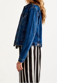 Femme portant une veste en denim bleue et une jupe rayée verticalement noir et blanc, vue de profil sur un fond uni.