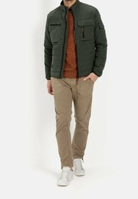 Quiltet grøn jakke med to brystlommer og lynlås. Båret over en tekstureret orange sweater, kombineret med beige bukser og hvide sneakers.